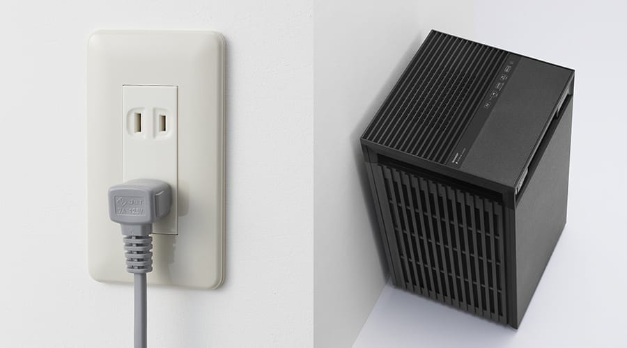 白い壁のコンセントに差し込まれたグレーのL字電源プラグと、壁際に置かれたFU-M1200イメージ。